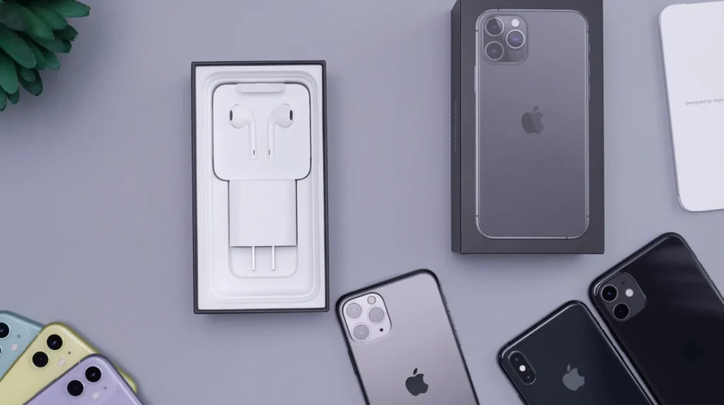 Flat arrangement of several iPhones and their accessories on a gray surface. An open iPhone case shows white earphones and charger, next to a closed iPhone case. Several iPhones are seen, including a gray one with triple camera, two black iPhones, and a group of colored iPhones (blue, yellow, purple) in the lower left corner.